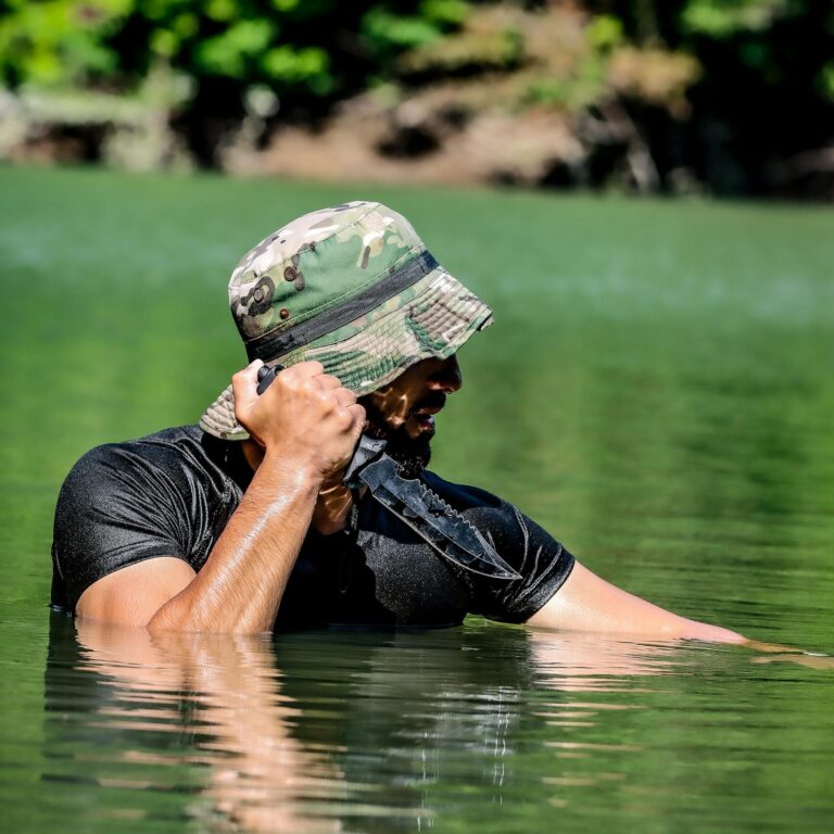 A man in camo hat holding a knife while standing in a river, portraying survival skills.